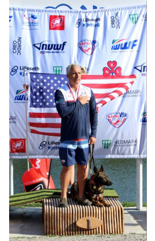 Mike Royal has hand over his heart and wears the USA Adaptive Water Ski Team uniform with gold medal hanging around his neck. He is standing on top of the podium during the audio slalom award presentation at the 2019 IWWF World Disabled Waterski Championships. Subi, his Fidelco German shepherd Guide Dogwears harness and lays next to Mike.