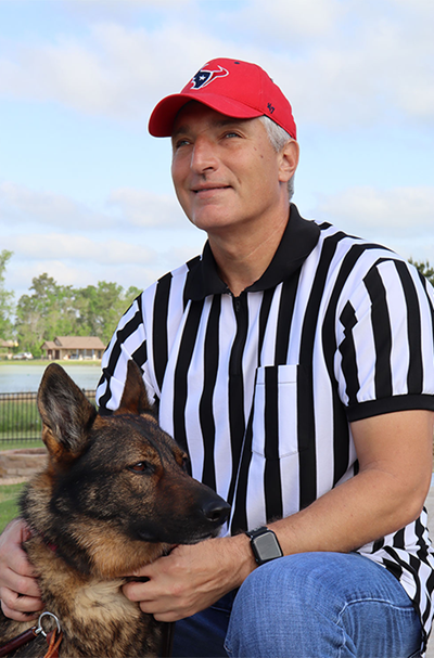 Mike Royal kneels next to his guide dog Subi. Mike is a middle-aged gray-haired man wearing a battle-red Houston Texan hat and a black-and-white striped referee uniform. Subi is a four-year-old sable-colored female German Shepherd wearing a Fidelco Guide Dog harness.